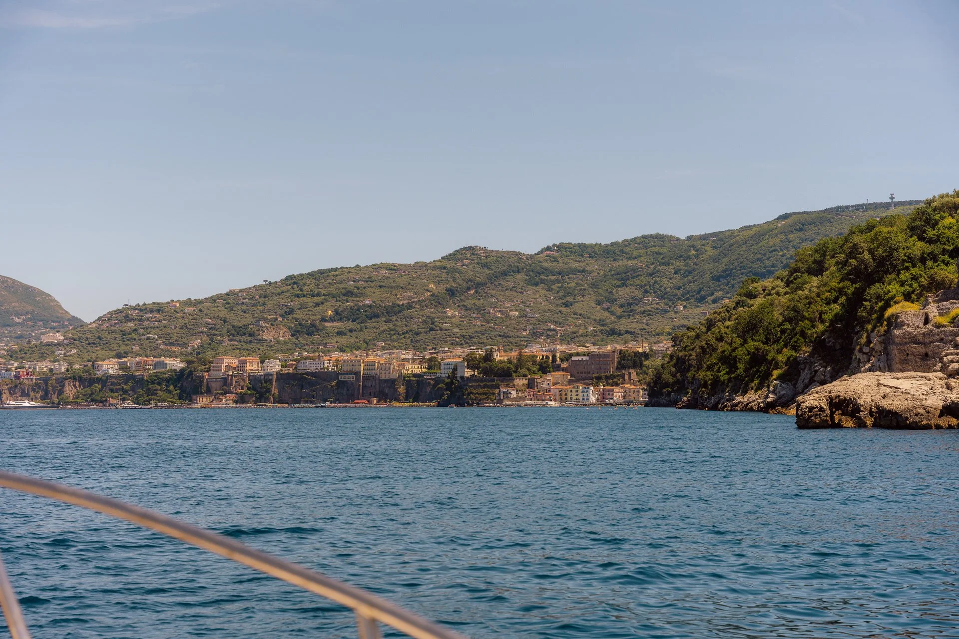 View of a coastline with buildings along the shore and lush green hills in the background, seen from a boat on the water.