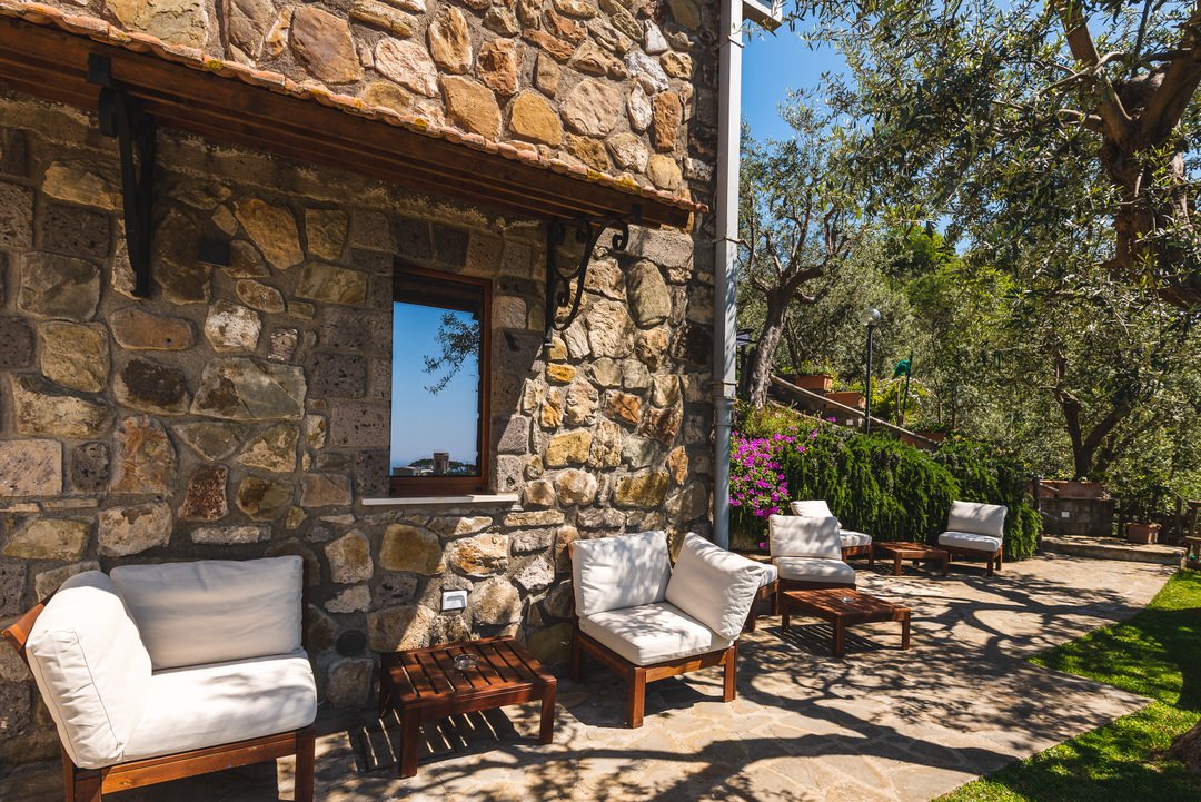 Outdoor patio area with stone wall, white cushioned wooden chairs, small tables, lush trees, and flowering bushes in bright sunlight.