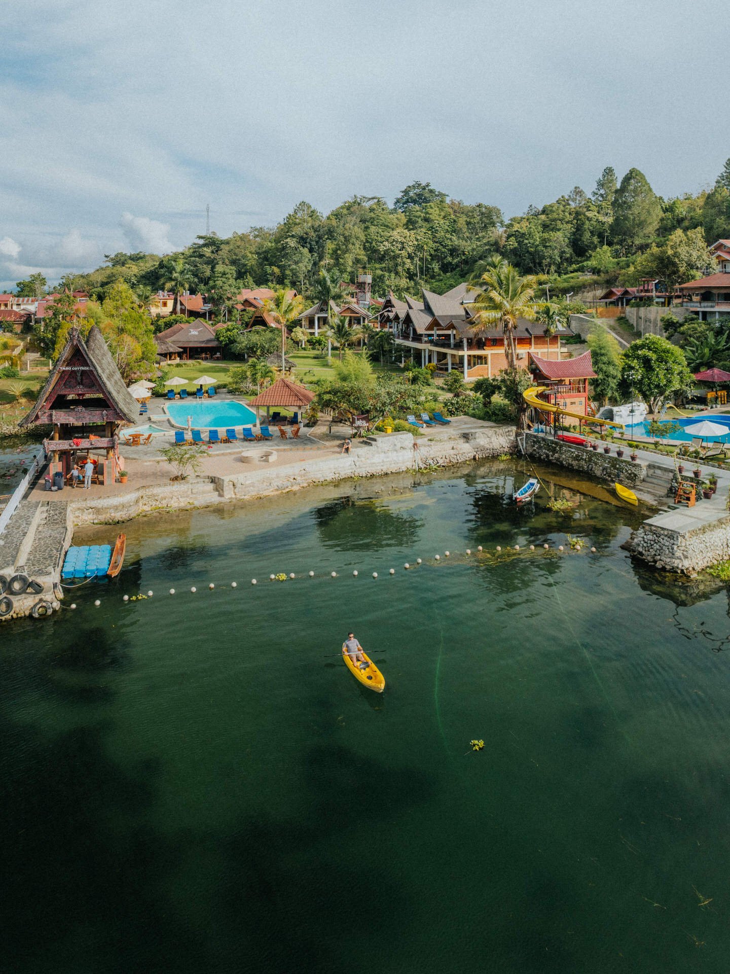 A tropical resort with a lake, swim area, pool, and houses surrounded by lush greenery. A person paddles a yellow kayak on the lake.