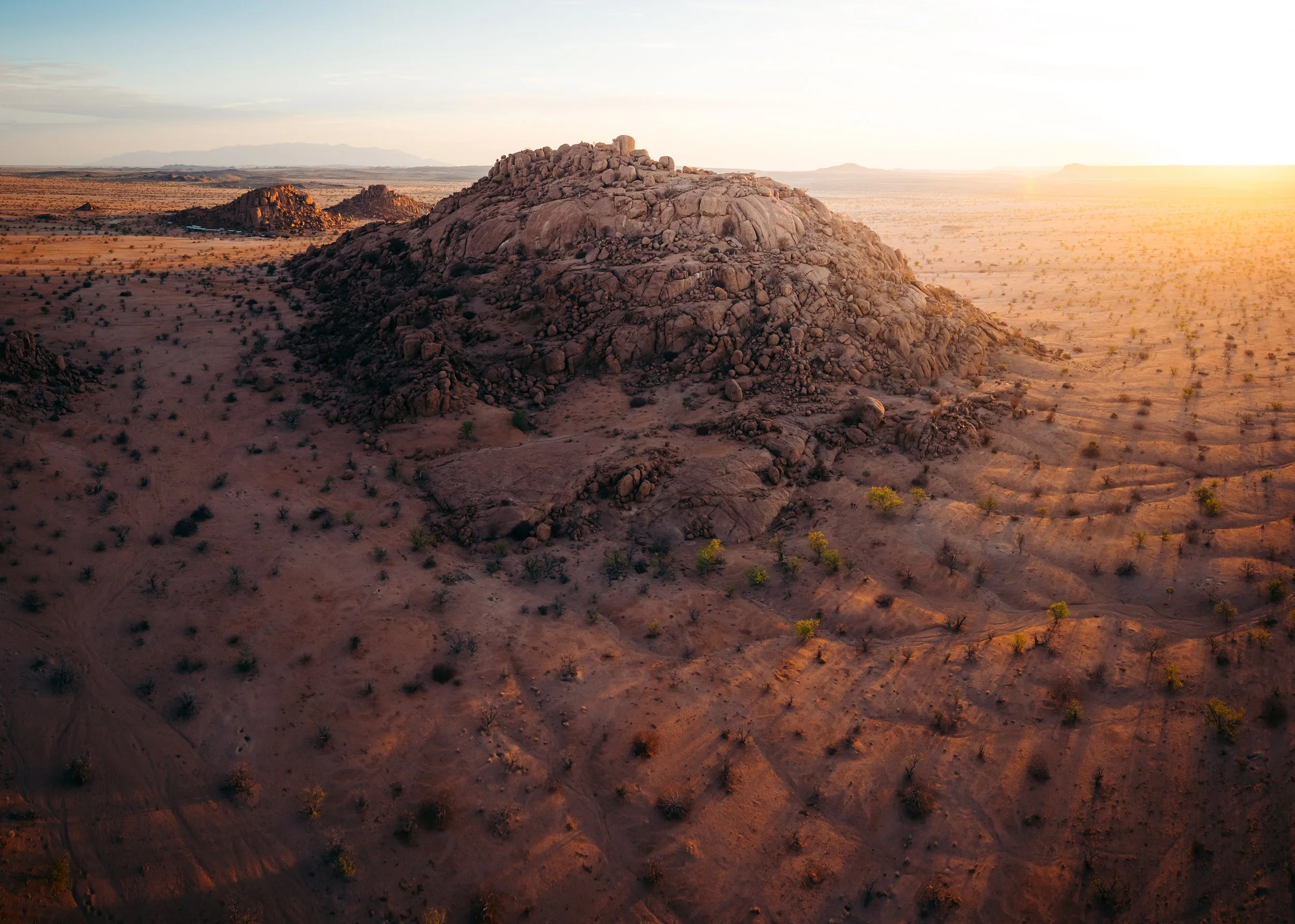 A desert landscape at sunset with a rocky hill in the center, sparse vegetation, and distant mountains on the horizon.