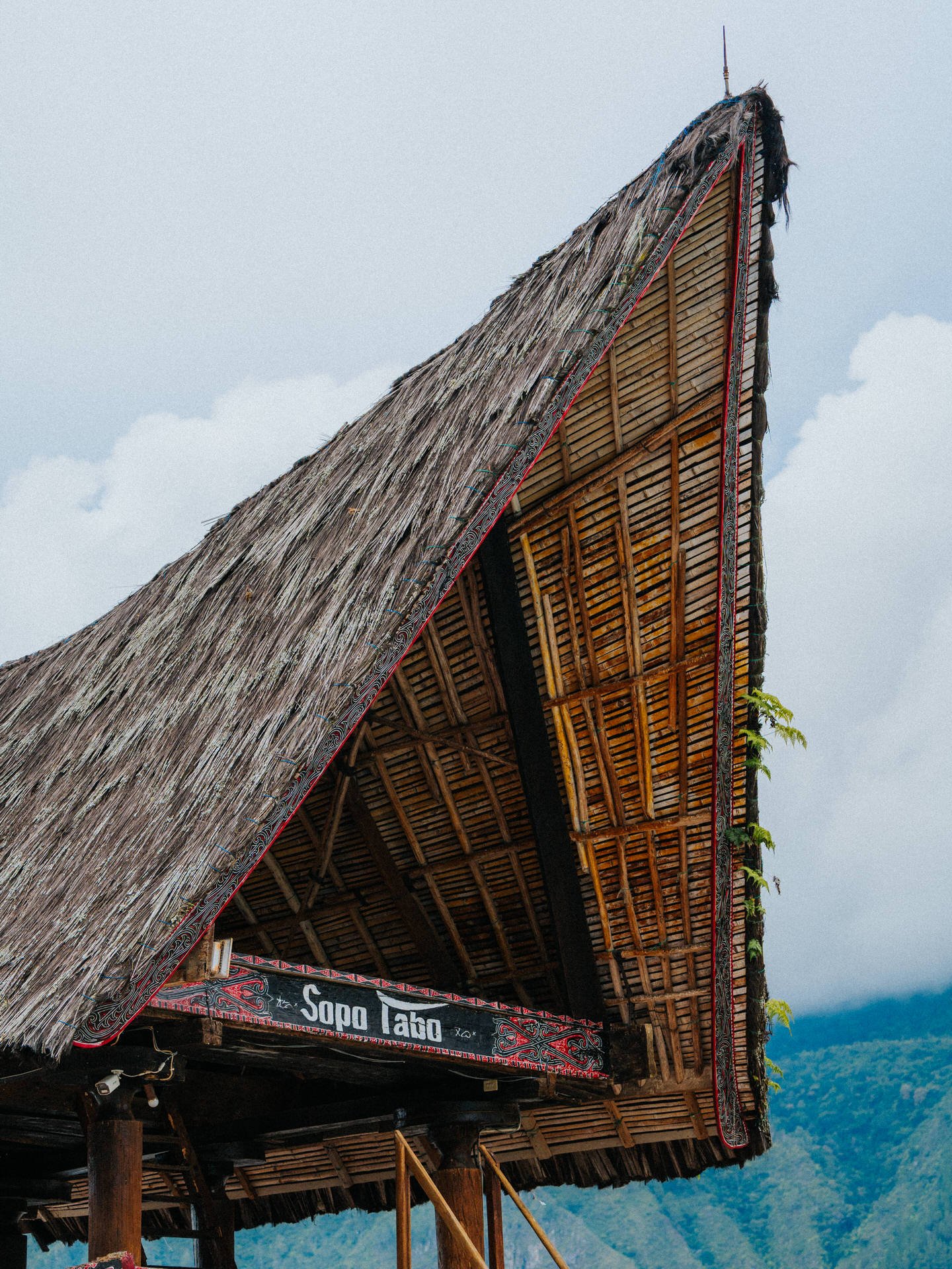 Thatched roof of a traditional building with a wooden sign that reads 'Sopo Tabo', set against a backdrop of mountains and cloudy sky.