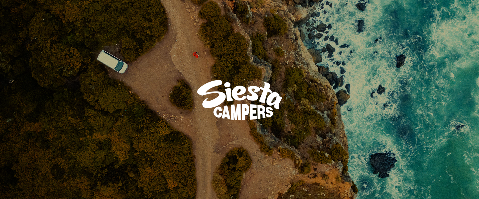 Aerial view of a camping site with a vehicle parked on a dirt path surrounded by bushes, overlooking the ocean with waves crashing on rocks.