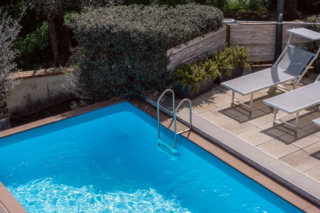 Above-ground swimming pool with a metal ladder next to a tiled patio area with lounge chairs and potted plants.