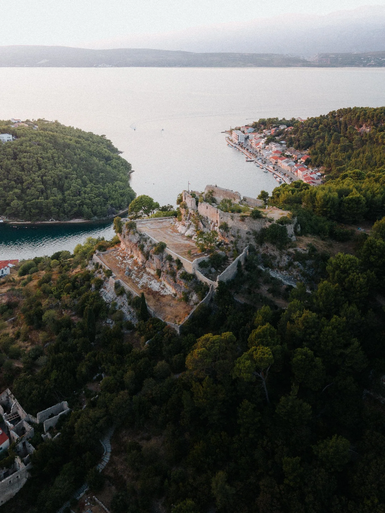 Aerial view of a coastline with dense green trees, old ruins on a hill, and a small town with colorful buildings along the water, with mountains in the background.