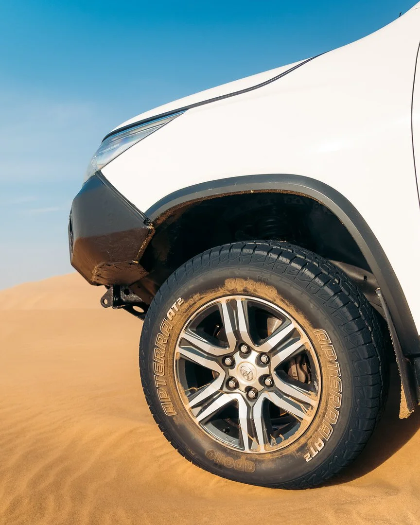Close-up of a white off-road vehicle's front wheel on sand dunes, showing a tire marked 'Aptie Terrain' and a black fender flaring around the wheel.