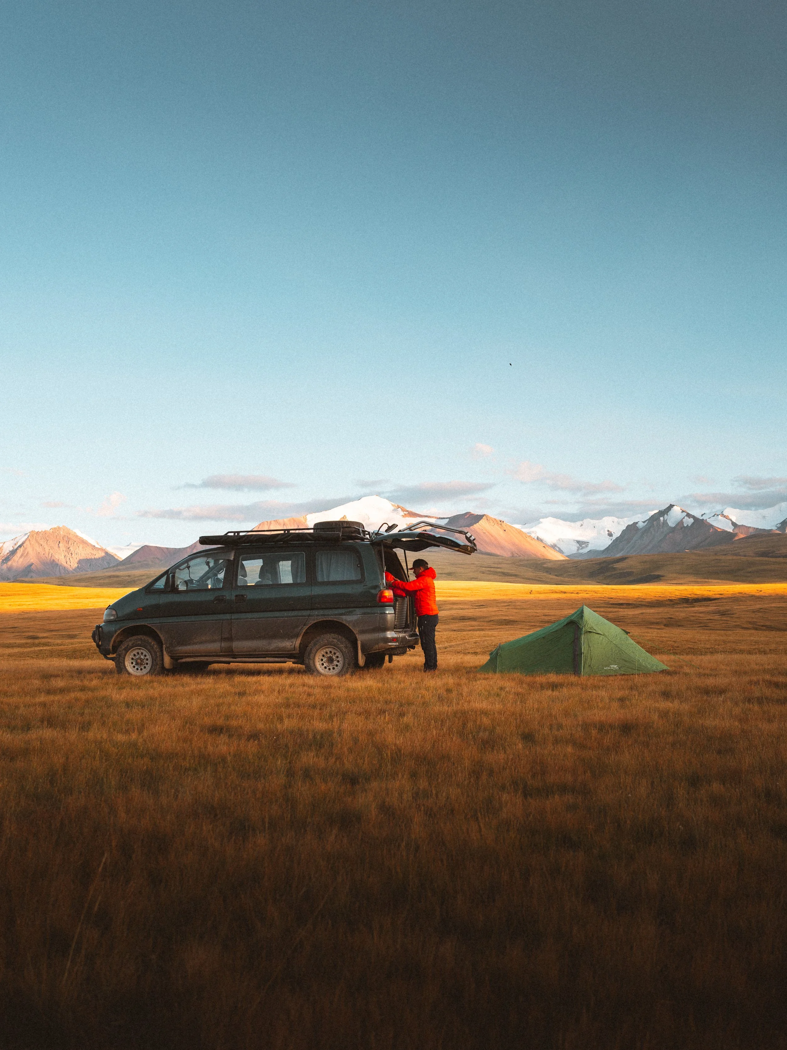 Person in a red jacket standing outside a black van with the rear hatch open, next to a green tent on an open grassy plain with mountains in the background during daytime.