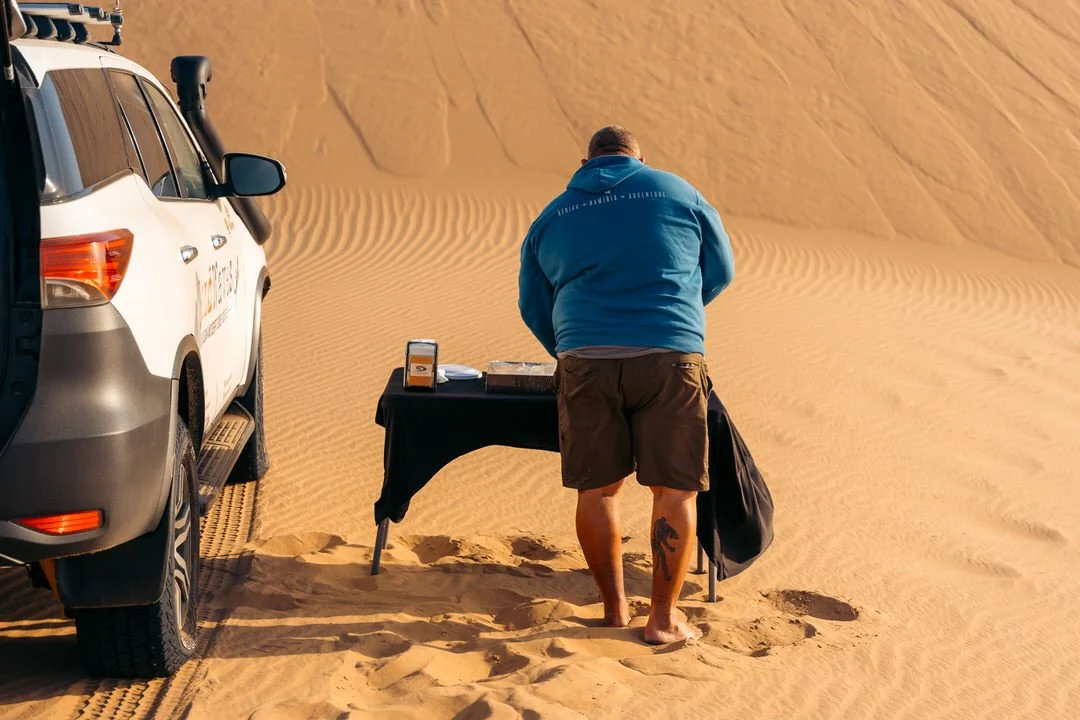 A man in a blue jacket and brown shorts standing behind a black table in a desert with sand dunes, next to a white off-road vehicle.