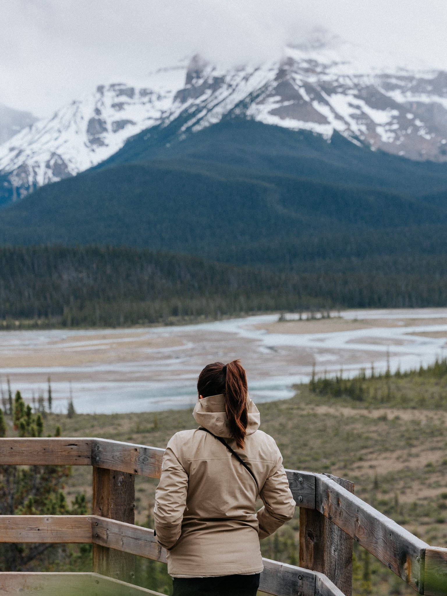 A woman with brown hair tied back wearing a beige jacket and black pants standing on a wooden observation deck overlooking a landscape of mountains with snow, green trees, and a river or stream in the foreground.