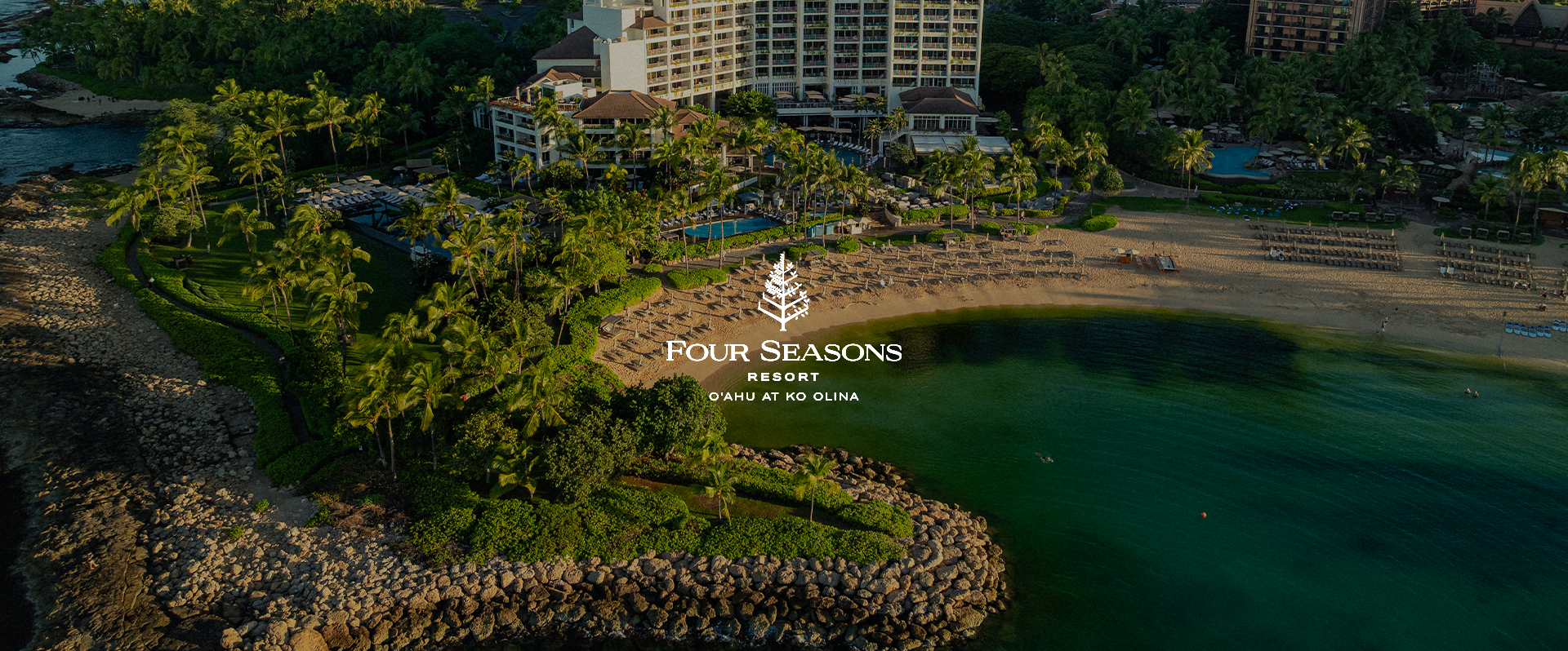 Aerial view of the Four Seasons Resort Oahu at Ko Olina, showing a beach with sun loungers, umbrellas, a resort building with multiple floors, a swimming pool with palm trees, and the ocean.