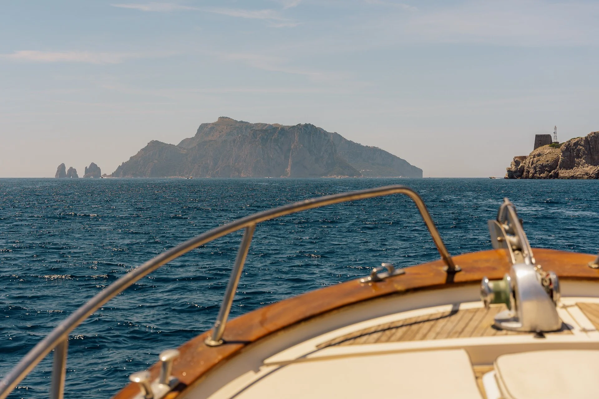 View from a boat showing open water and a distant island with steep cliffs and rock formations, under a partly cloudy sky.