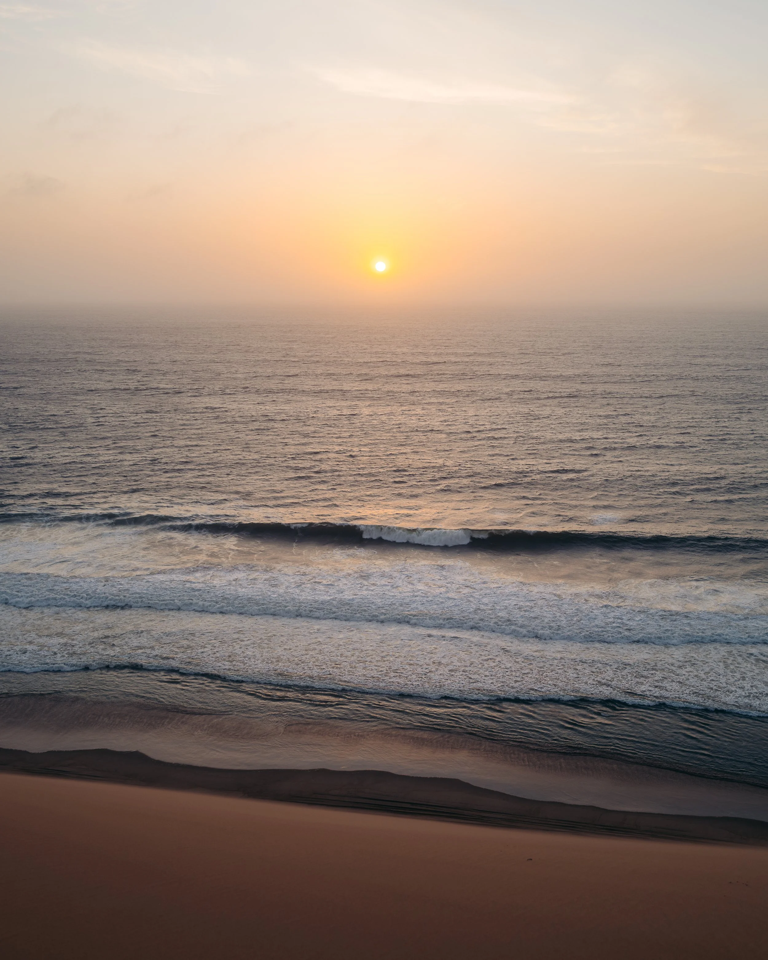 Sunset over the ocean with waves hitting the sandy beach in the foreground.