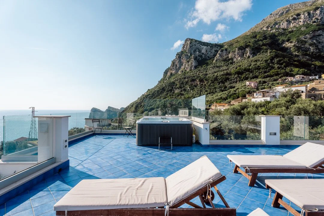 Rooftop terrace with blue tiled floor, lounge chairs, hot tub, glass railing, overlooking green mountains and ocean under a clear sky.
