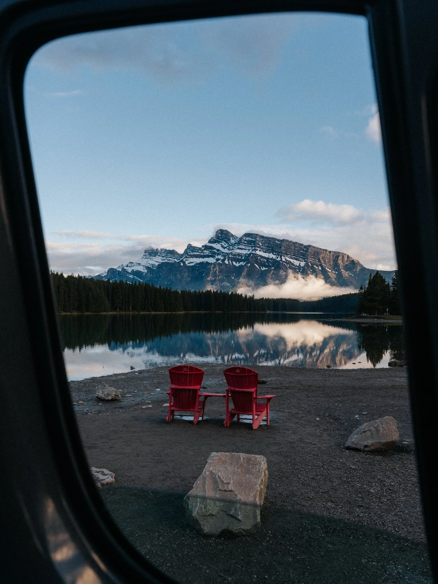 Two red Adirondack chairs on a black gravel beach facing a calm lake, with a mountain range in the background and a partly cloudy sky.