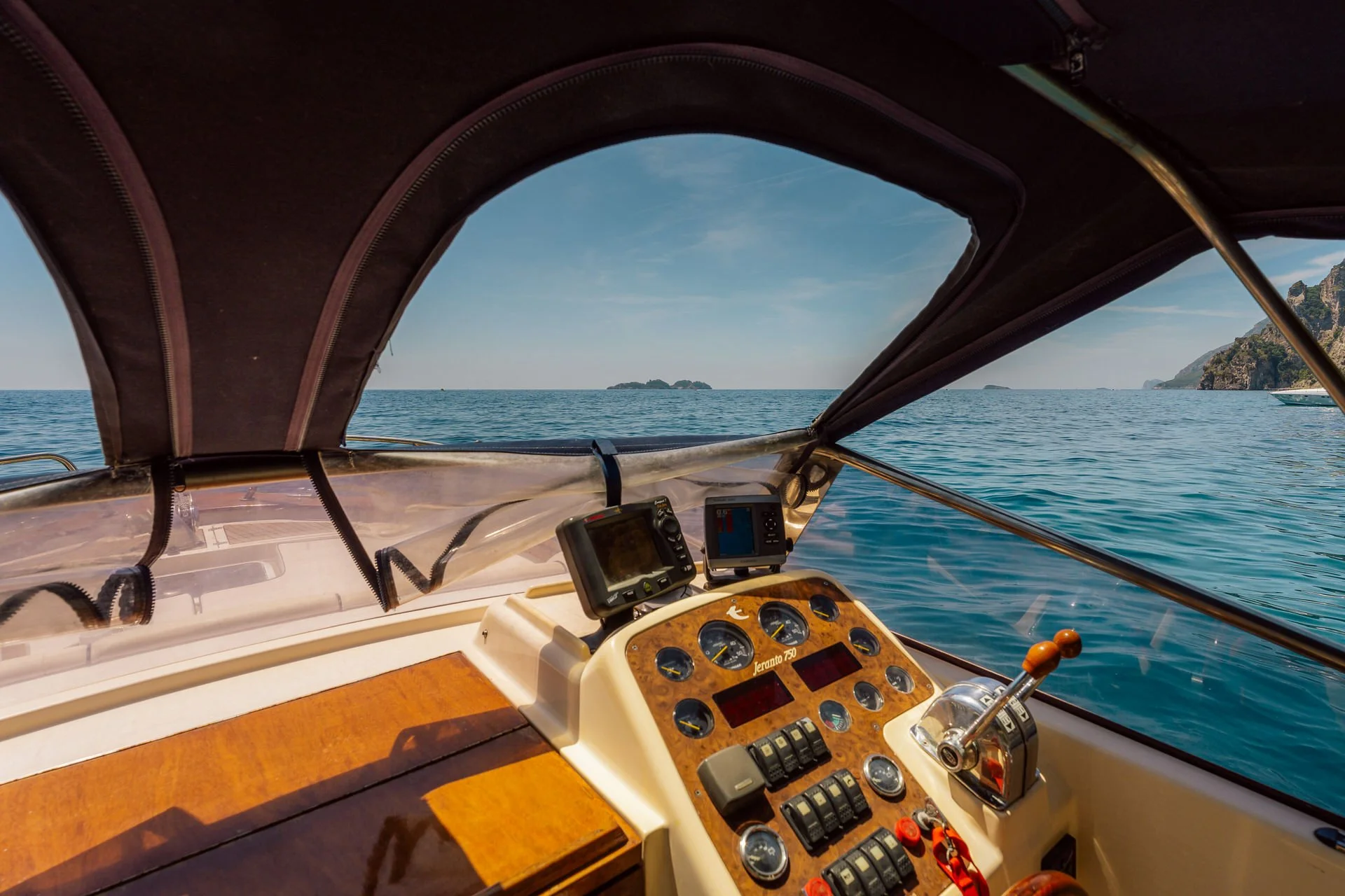 Inside view of a boat cockpit showing navigation and control instruments, with a view of the ocean and distant islands under a partly cloudy sky.