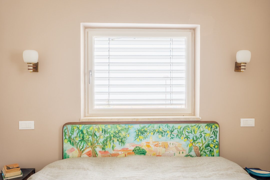 View of a bedroom with a window, beige walls, two wall sconces, and a colorful headboard painted with trees and buildings.