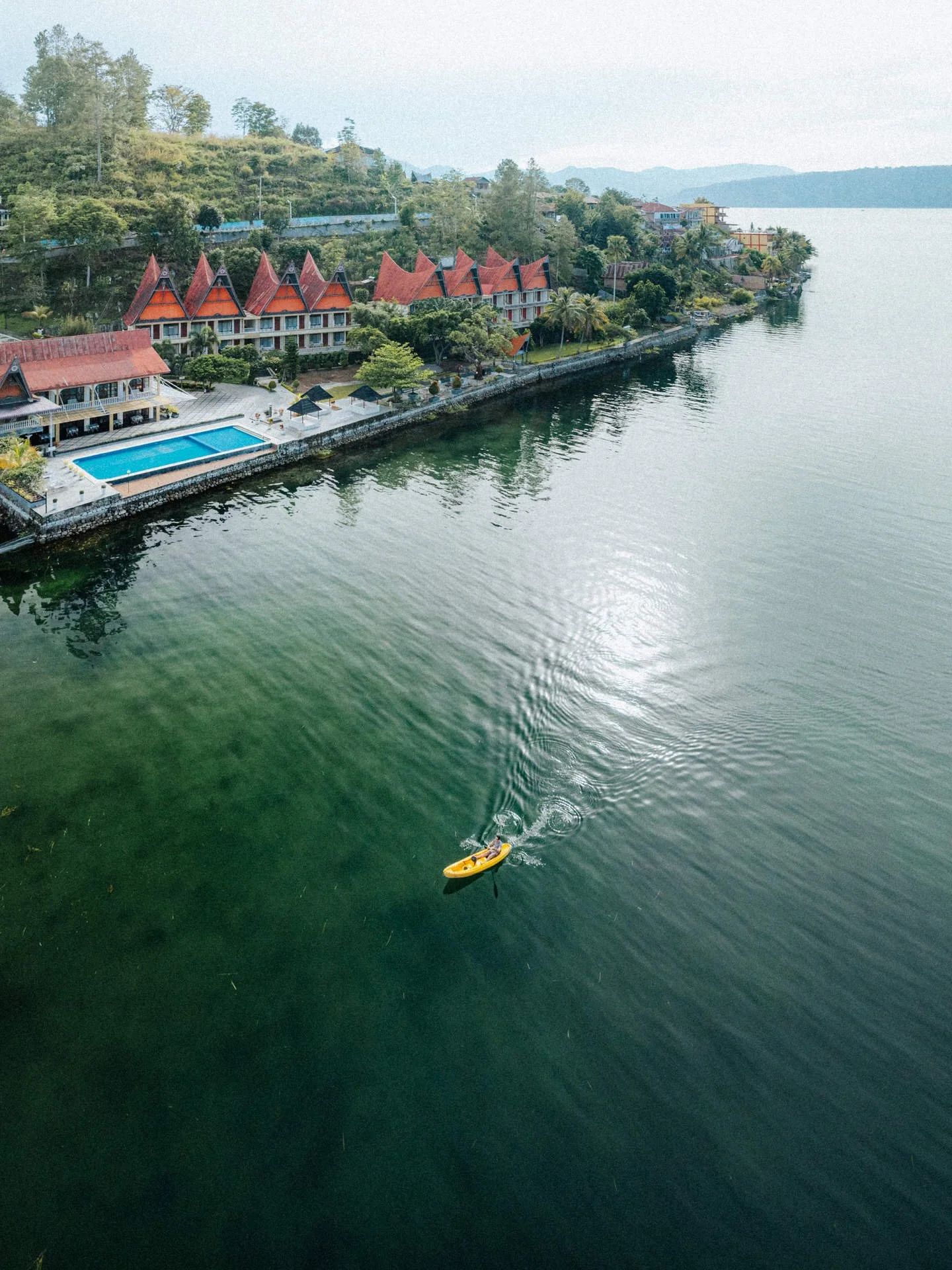Aerial view of a lakeside resort with traditional-style buildings, a swimming pool, and a person kayaking on the water.