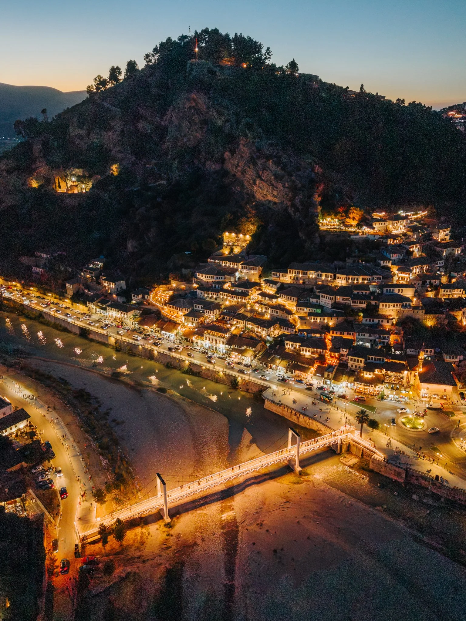 Aerial night view of a city with a river and a bridge, buildings lit up along the water, and a mountain with trees and structures illuminated on top.