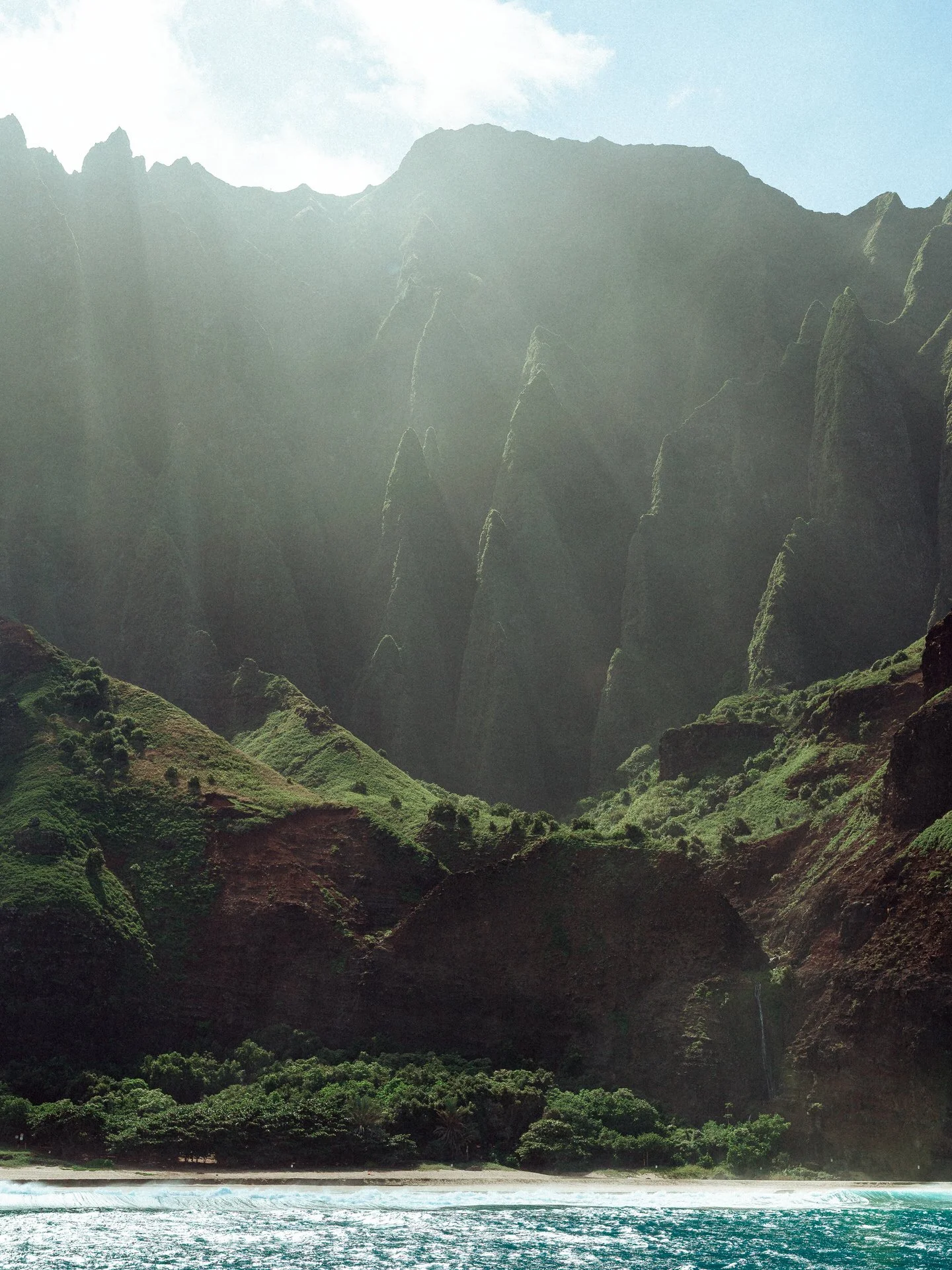 Tropical island with green mountains towering over a sandy beach, blue ocean at shoreline, and sunlight filtering through the sky.