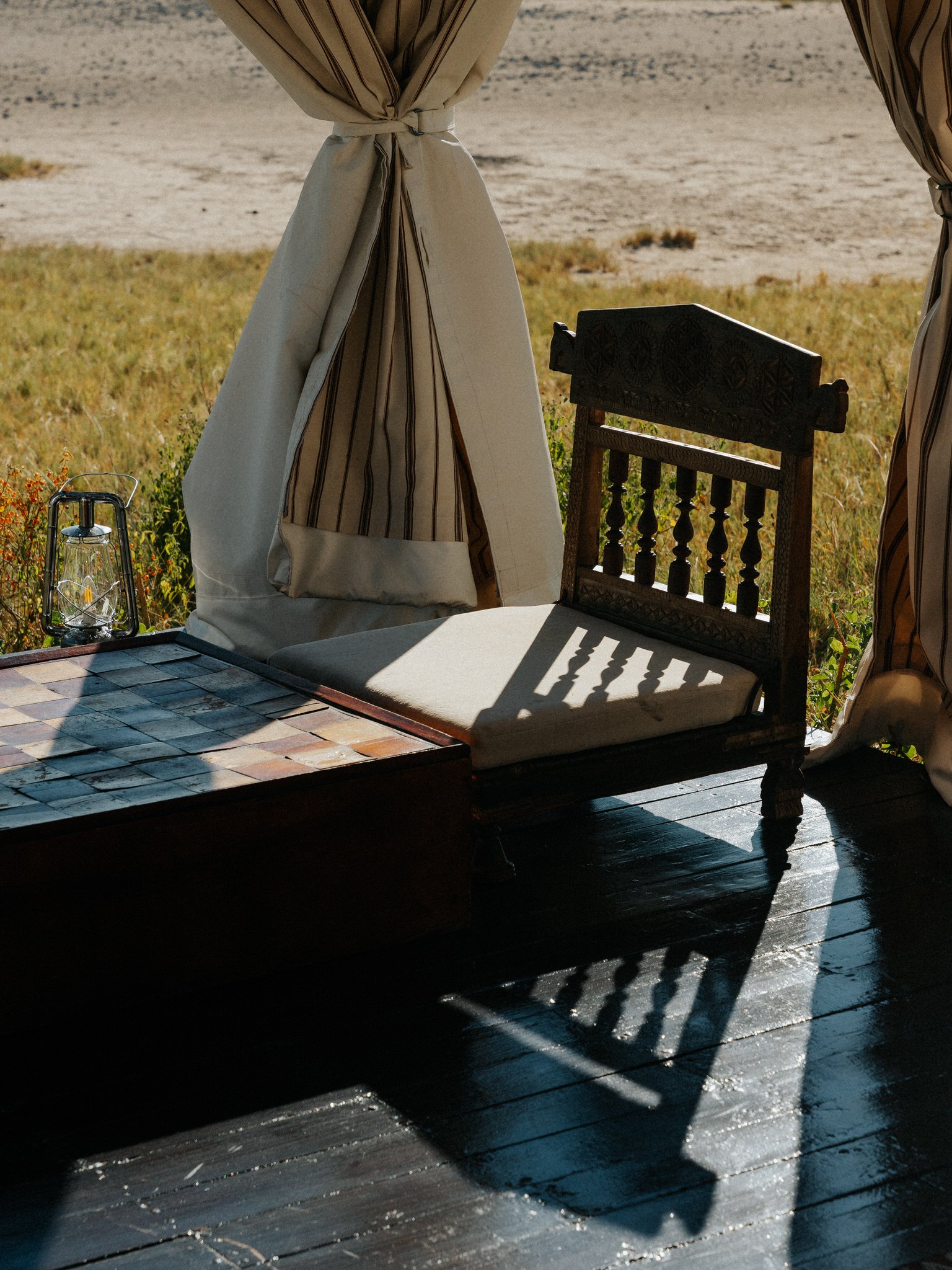 A wooden table with a tiled surface, partly in shadow, with a clear glass lantern and a cushioned wooden chair on a porch overlooking a barren field in the background.