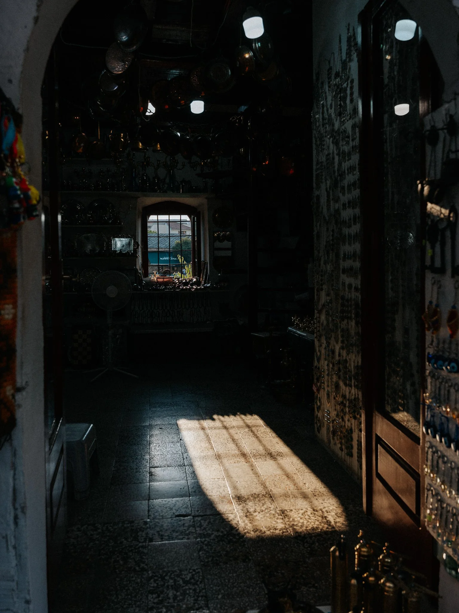 Dark interior room with sunlight streaming through a window, casting shadows on the floor. Shelves and various hanging objects are visible, creating a cozy, rustic atmosphere.
