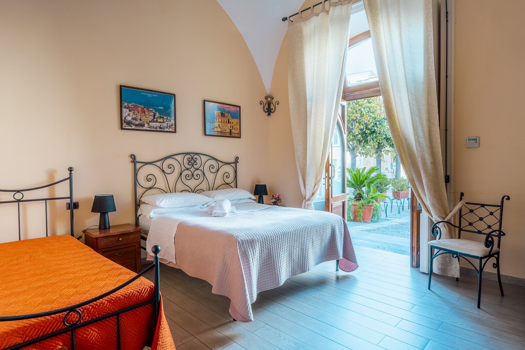 A bright hotel room with a double bed, orange single bed, and patio door with open curtains revealing outside greenery and patio furniture.