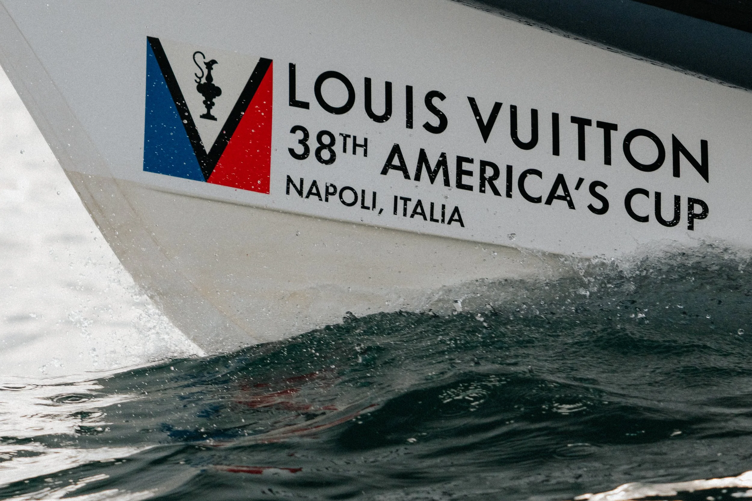 A boat with a Louis Vuitton logo, 38th America's Cup, Naples, Italy, written on the side, seen through water droplets and ripples on the water's surface.