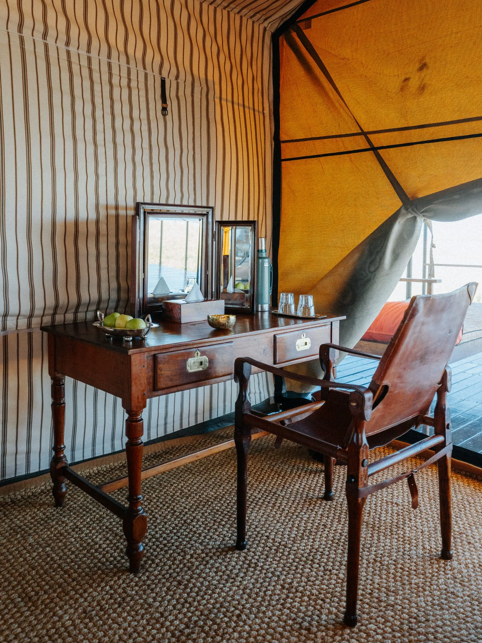 A wooden desk with a mirror, a thermos, and a bowl of green apples, with an armchair nearby, in a room with striped and solid yellow walls and a patterned carpet.