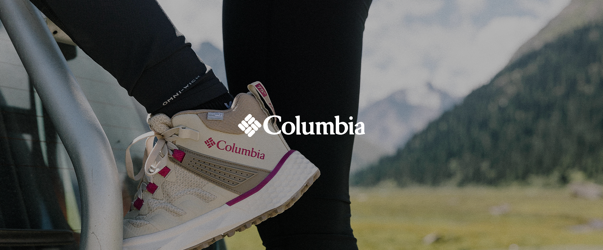 Close-up of a person's foot wearing a Columbia athletic shoe with beige, purple, and pink details, resting on a metal staircase railing outdoors with mountains and greenery in the background.