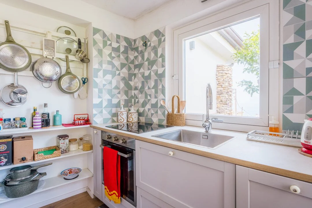 Bright kitchen with geometric backsplash, open shelving with pots, pans, and jars, window above sink, and countertop with dish rack and soap dispenser.