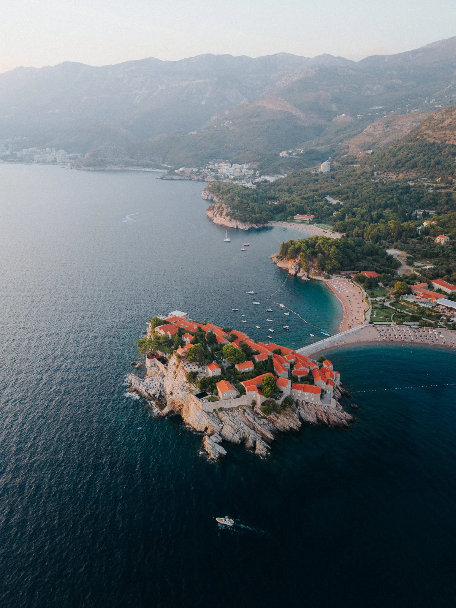 Aerial view of a coastal village on a rocky promontory with red-roofed buildings, a beach, and boats, surrounded by green hills and mountains.