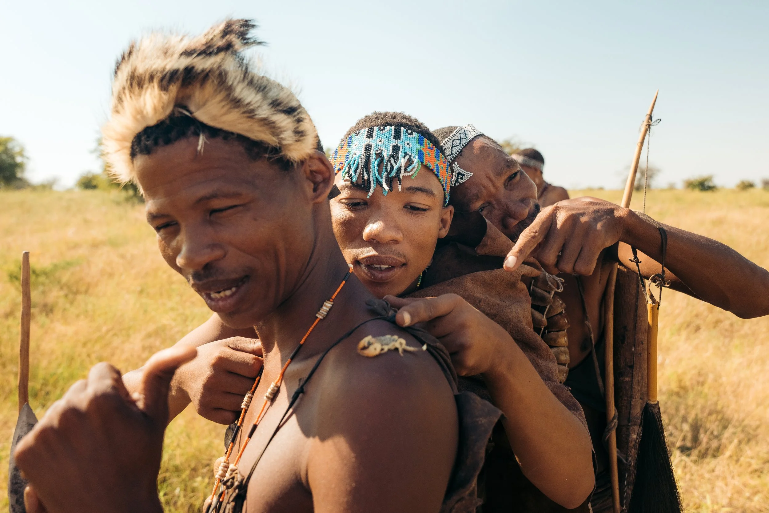 Group of indigenous people outdoors, wearing traditional beaded headpieces and necklaces, standing in a line with arms around each other's shoulders, in a grassy field under a clear sky.