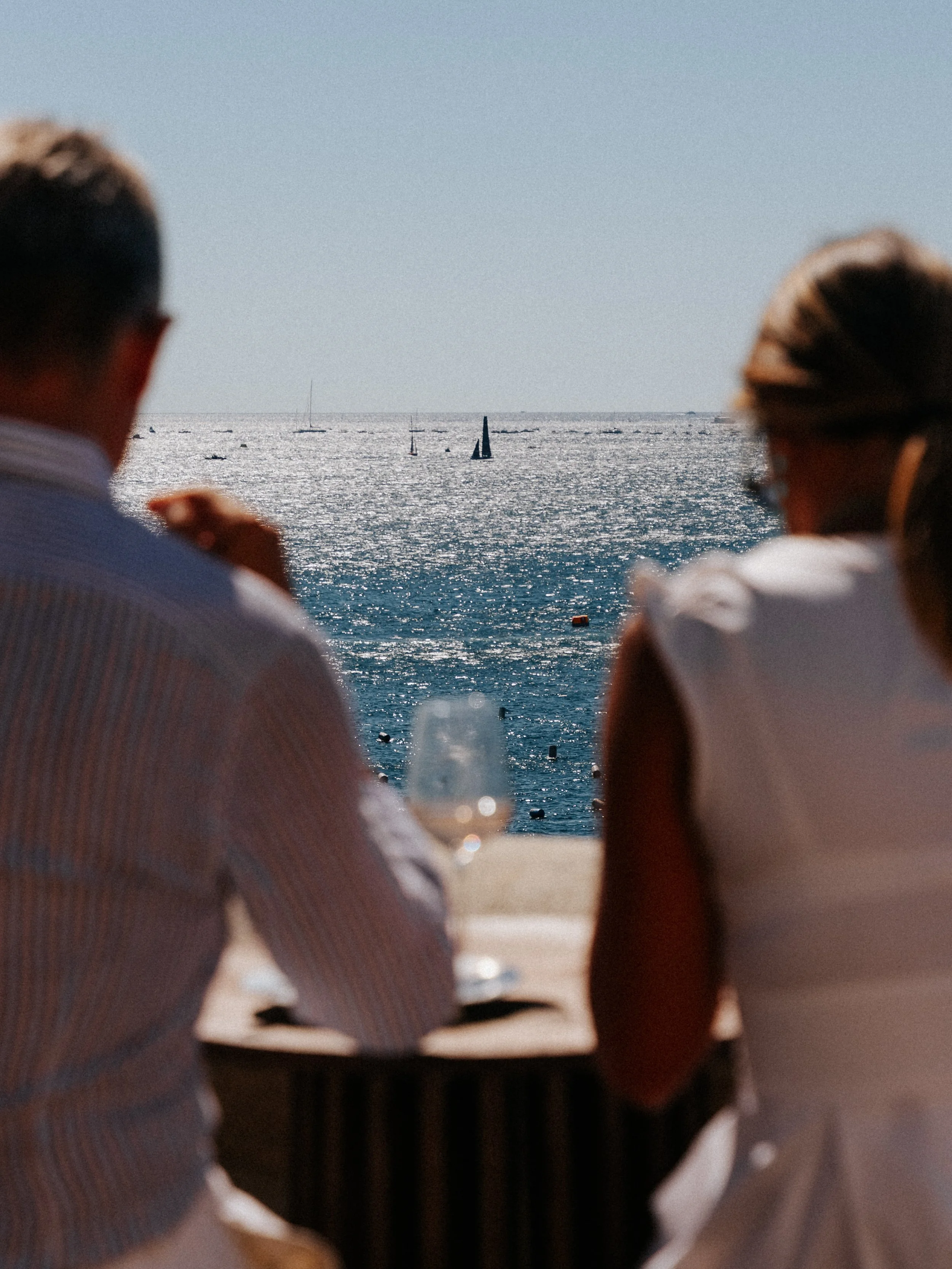 Two people sitting on the beach, looking at the ocean with sailboats in the distance.