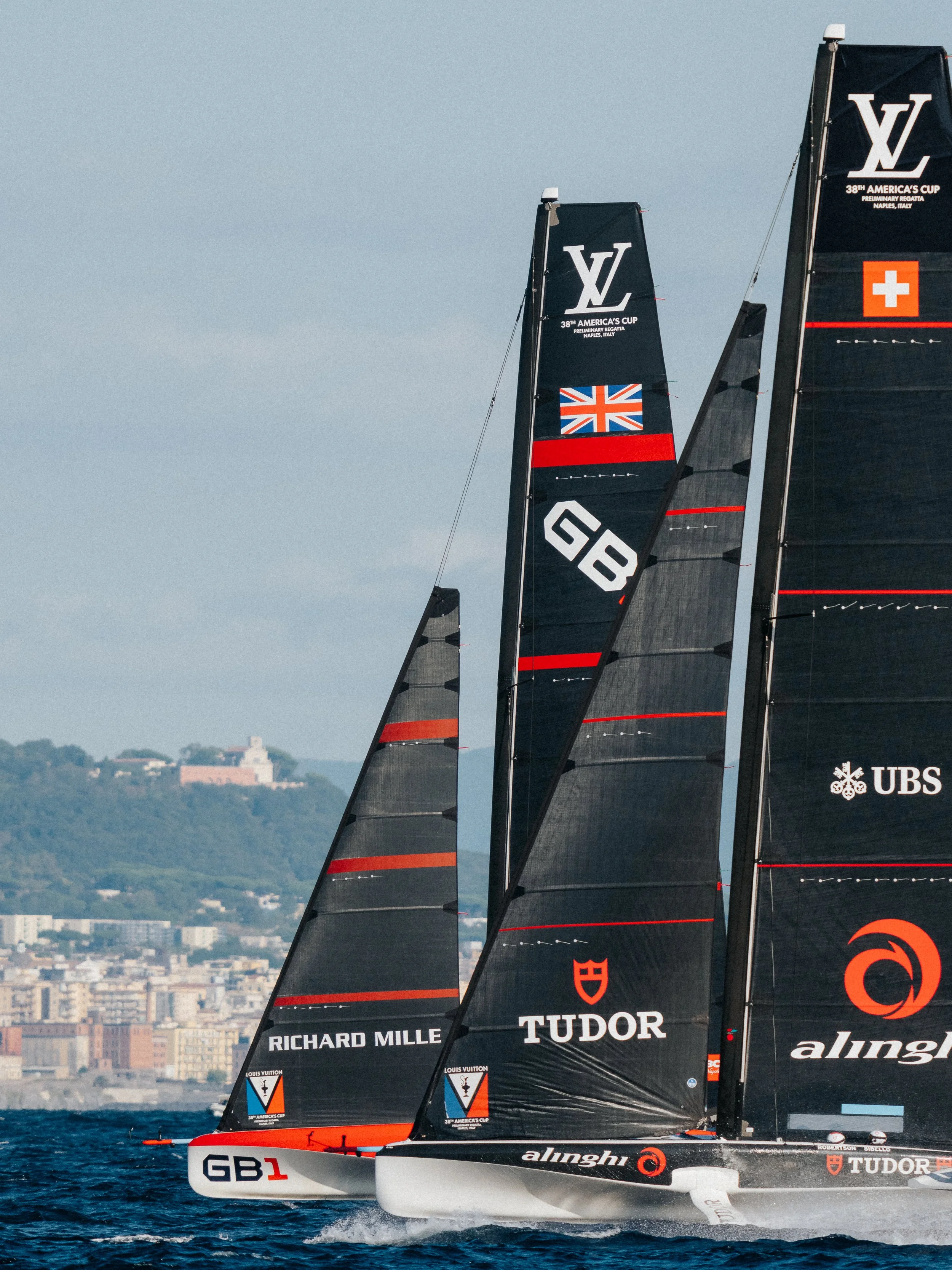 Yachting sailboats with black sails branded with sponsors' logos, racing in a regatta, with a cityscape and hills in the background.