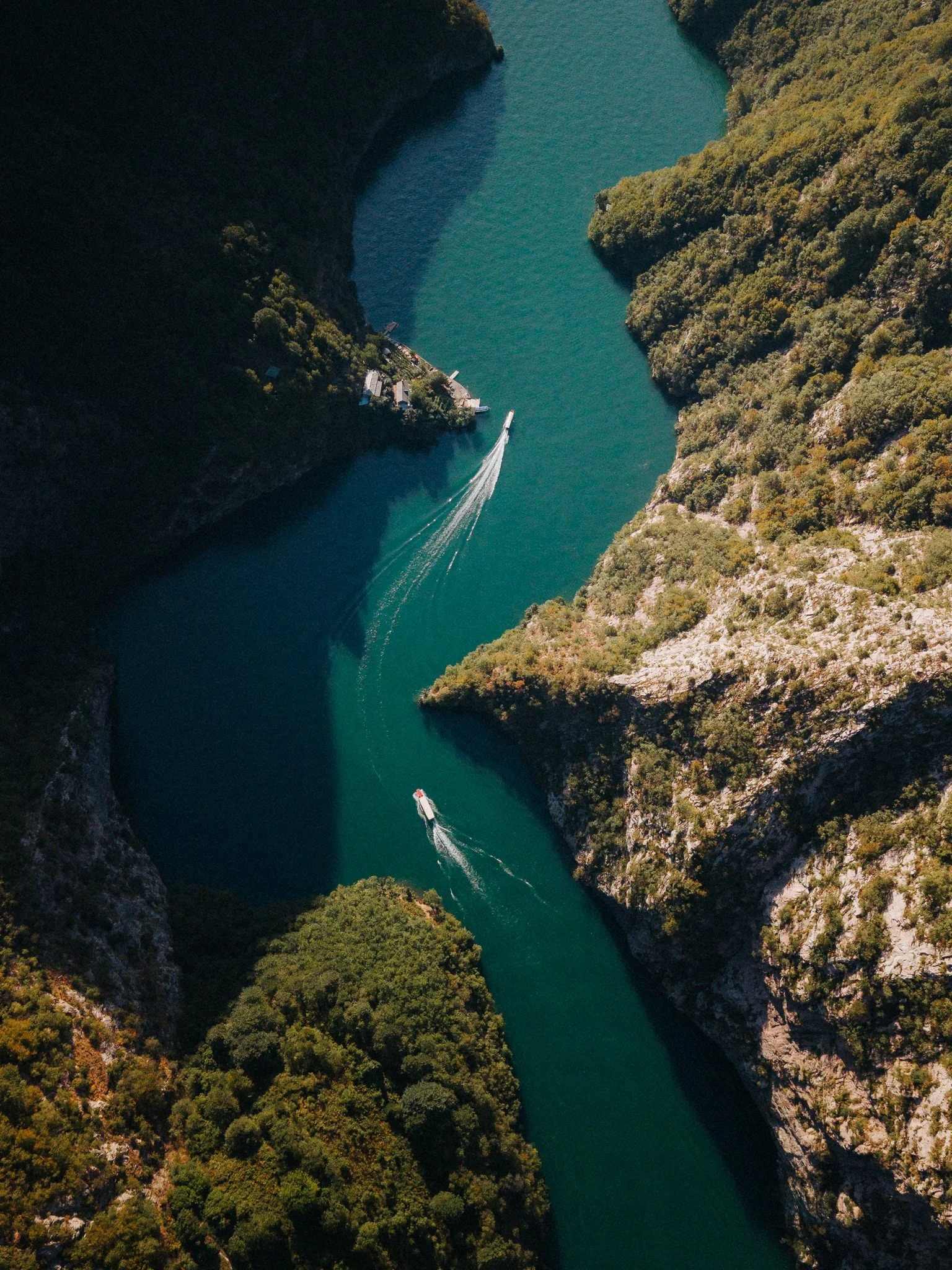 An aerial view of a narrow river flanked by lush green cliffs, with a boat speeding through the water creating a wake.