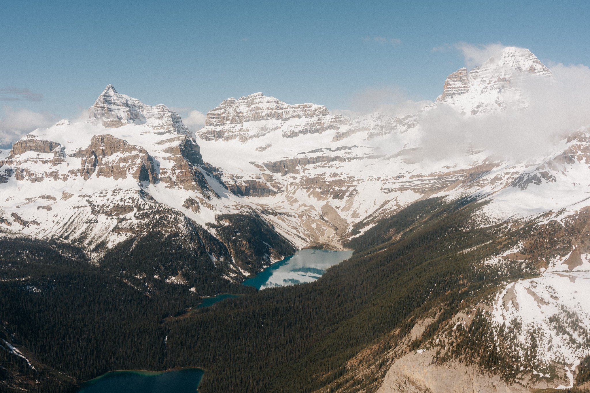 Snow-covered mountain range with a clear blue lake at the base, dense forested area, and partly cloudy sky.