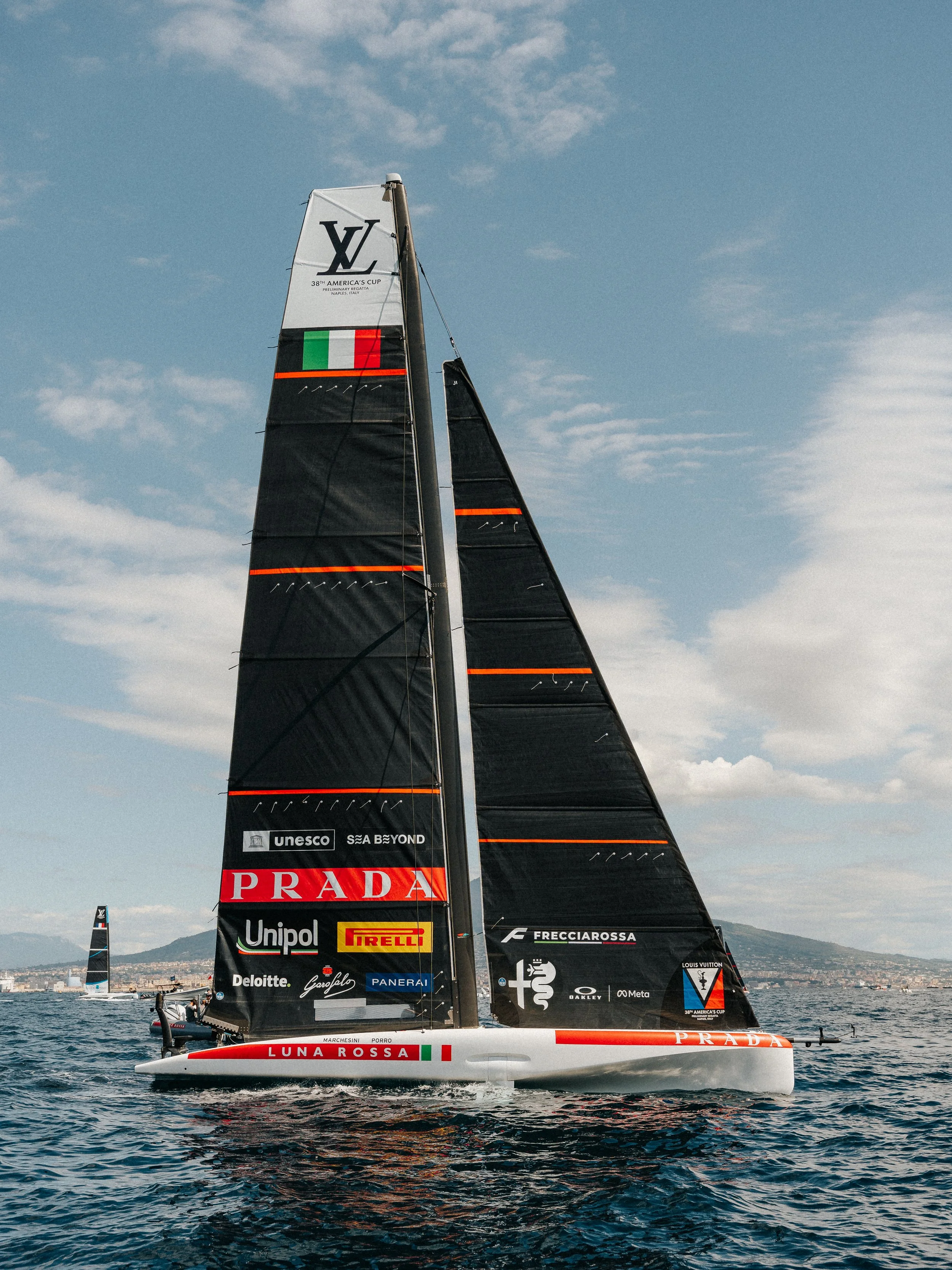 A racing sailboat with black sails featuring sponsor logos, including Prada, Luna Rossa, and Pirelli, competing on a body of water under a partly cloudy sky.