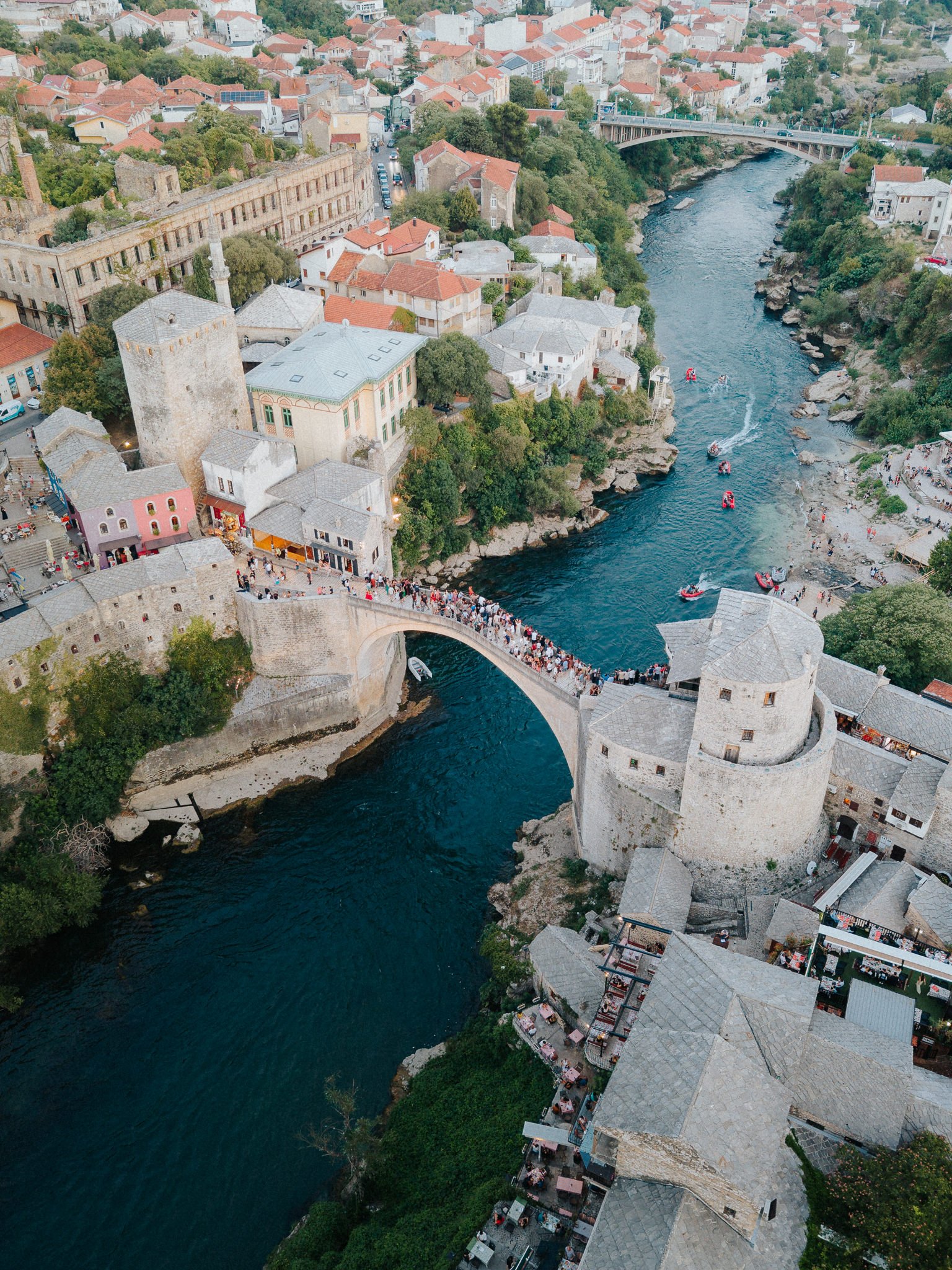 Aerial view of a historic European city with a river running through it. The river is crossed by a stone arch bridge crowded with people. On one side of the river, there are old stone buildings and towers, including a castle, mixed with modern struct
