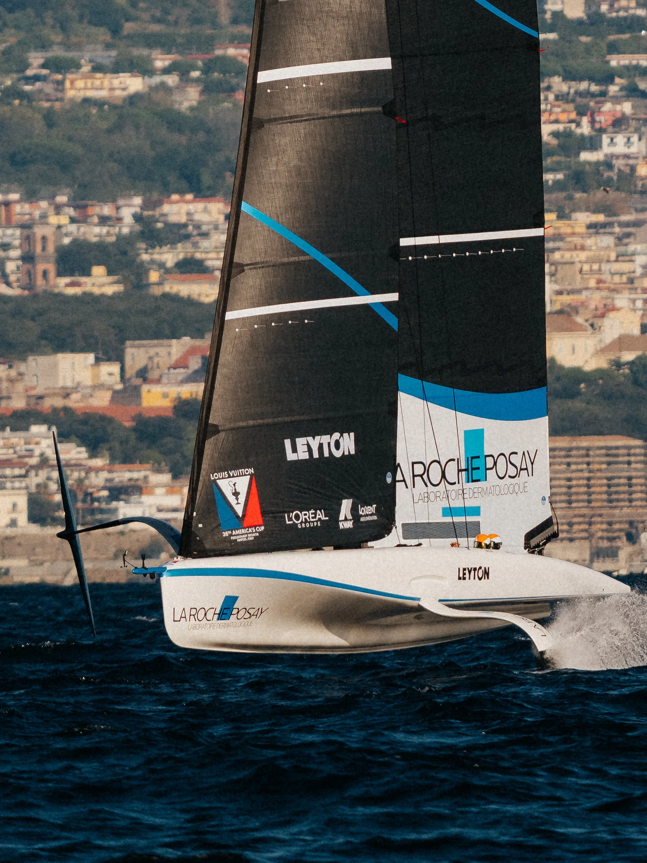 A racing sailboat on the water with a cityscape in the background, featuring black and white sails with blue accents, and logos including La Roche-Posay and Leyton.