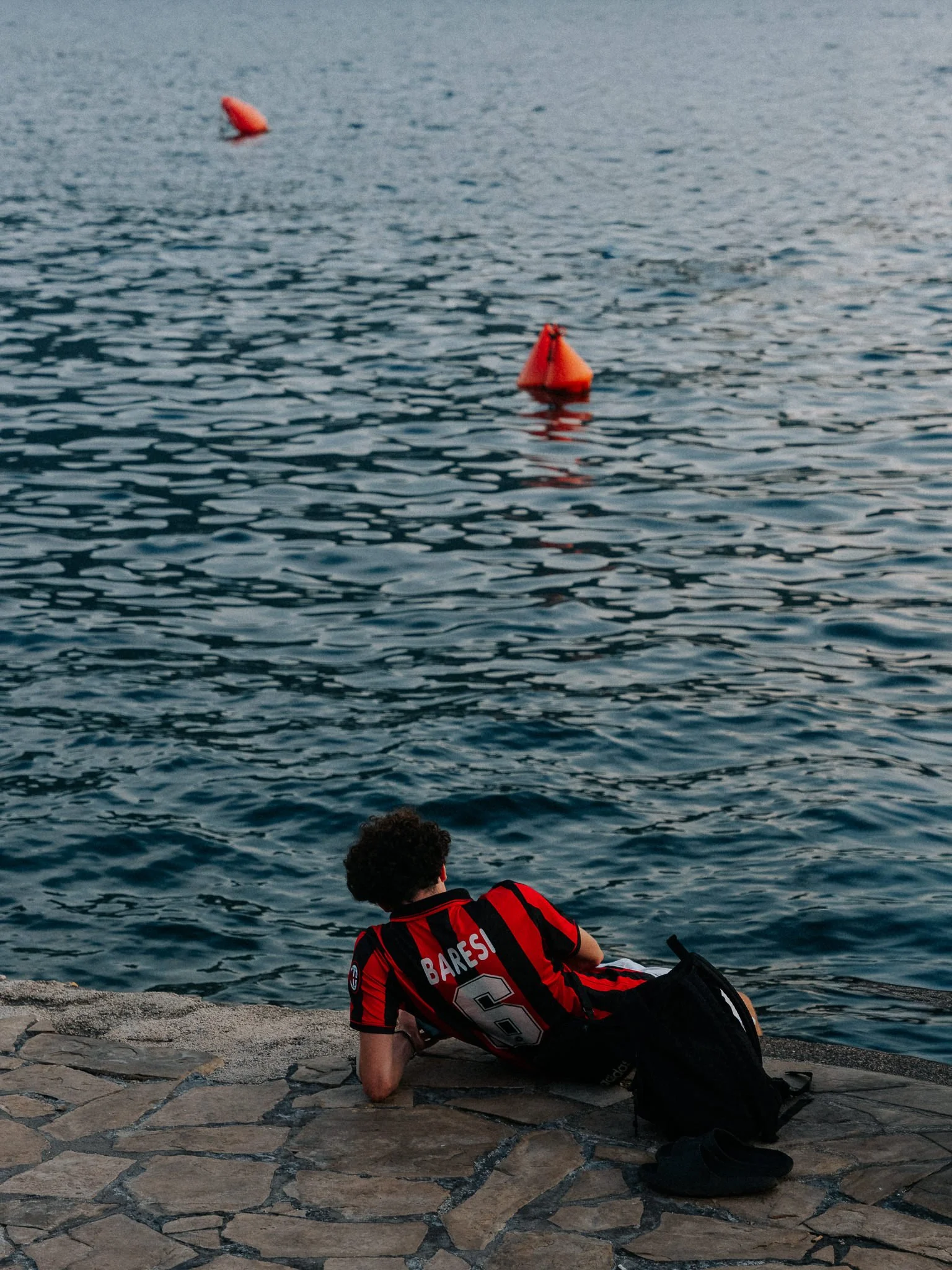 A person wearing a red and black sports jersey with the name 'Baresi' and the number 6, lying on a stone pier by the water, with legs crossed and head resting on one arm, facing away from the camera, and with buoys floating in the water.