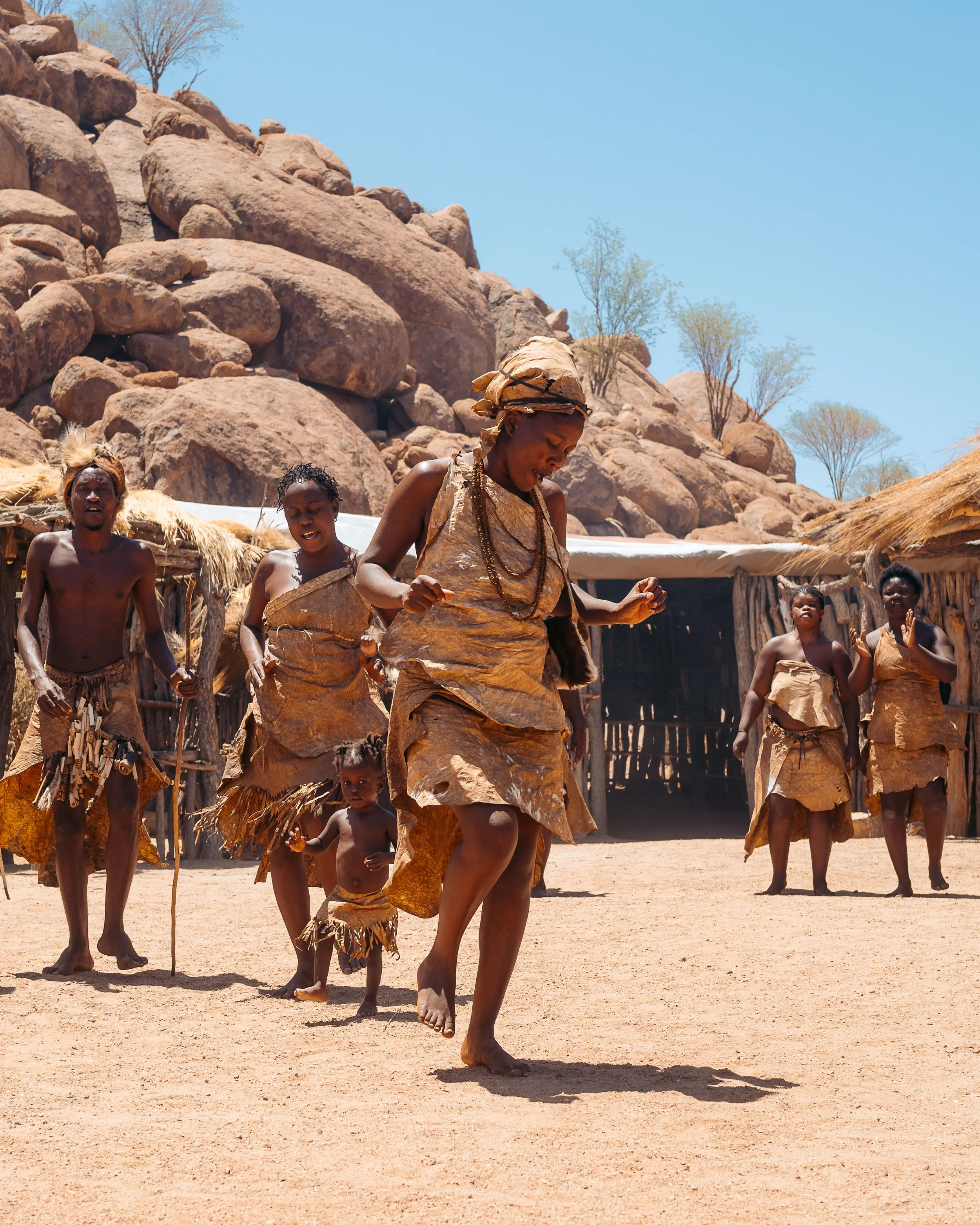 A group of people in traditional clothing dancing in a village with thatched huts, rocky hills, and sparse trees in the background under a clear blue sky.