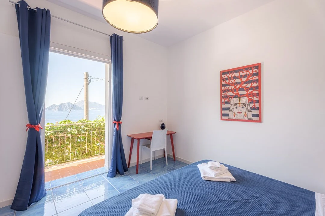A bedroom with a window view of the ocean and hills, blue curtains, a small desk with a white chair, a picture of a woman with a red tree on the wall, and a bed with folded towels.