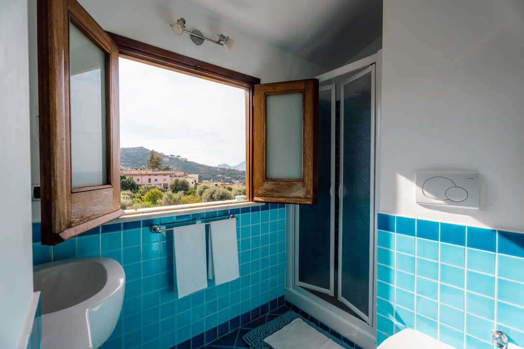 Bathroom with a wooden window open, blue tiled walls, a toilet, and a shower with a sliding door, overlooking a landscape with hills and buildings.
