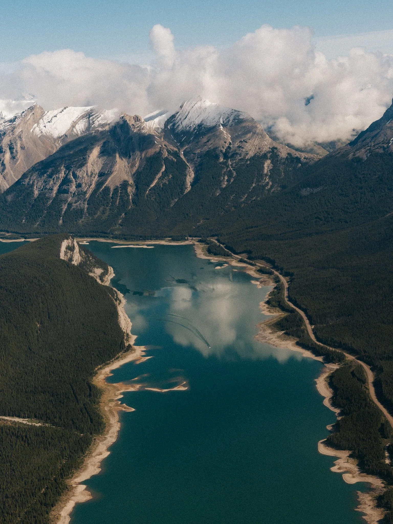 A large mountain range with snow-capped peaks partially covered by clouds, next to a wide river or lake surrounded by dense green forest, with a boat creating ripples in the water.