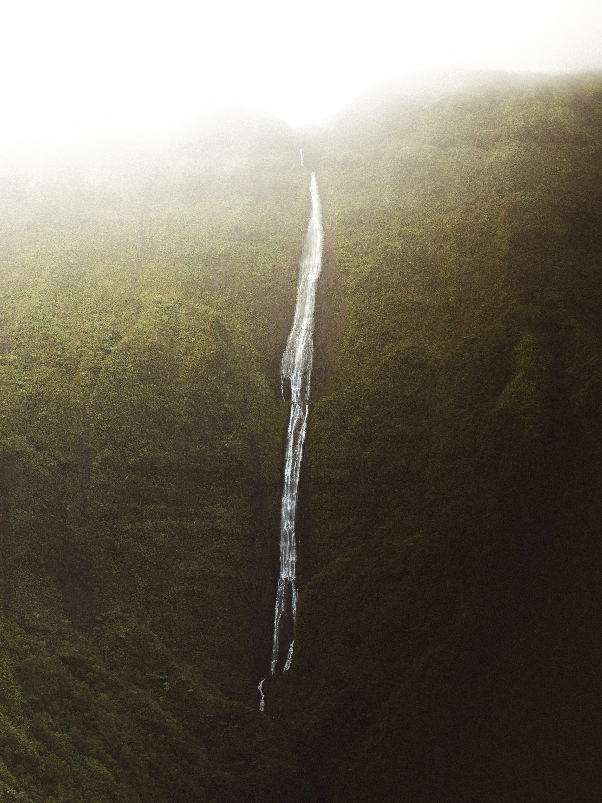 Tall waterfall cascading down green, moss-covered mountain slopes with mist at the top.