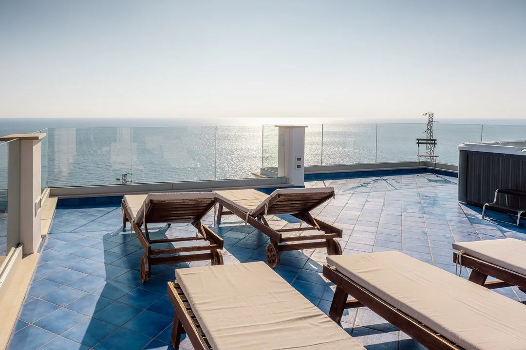 Rooftop terrace with wooden lounge chairs and a glass railing overlooking the ocean on a sunny day.