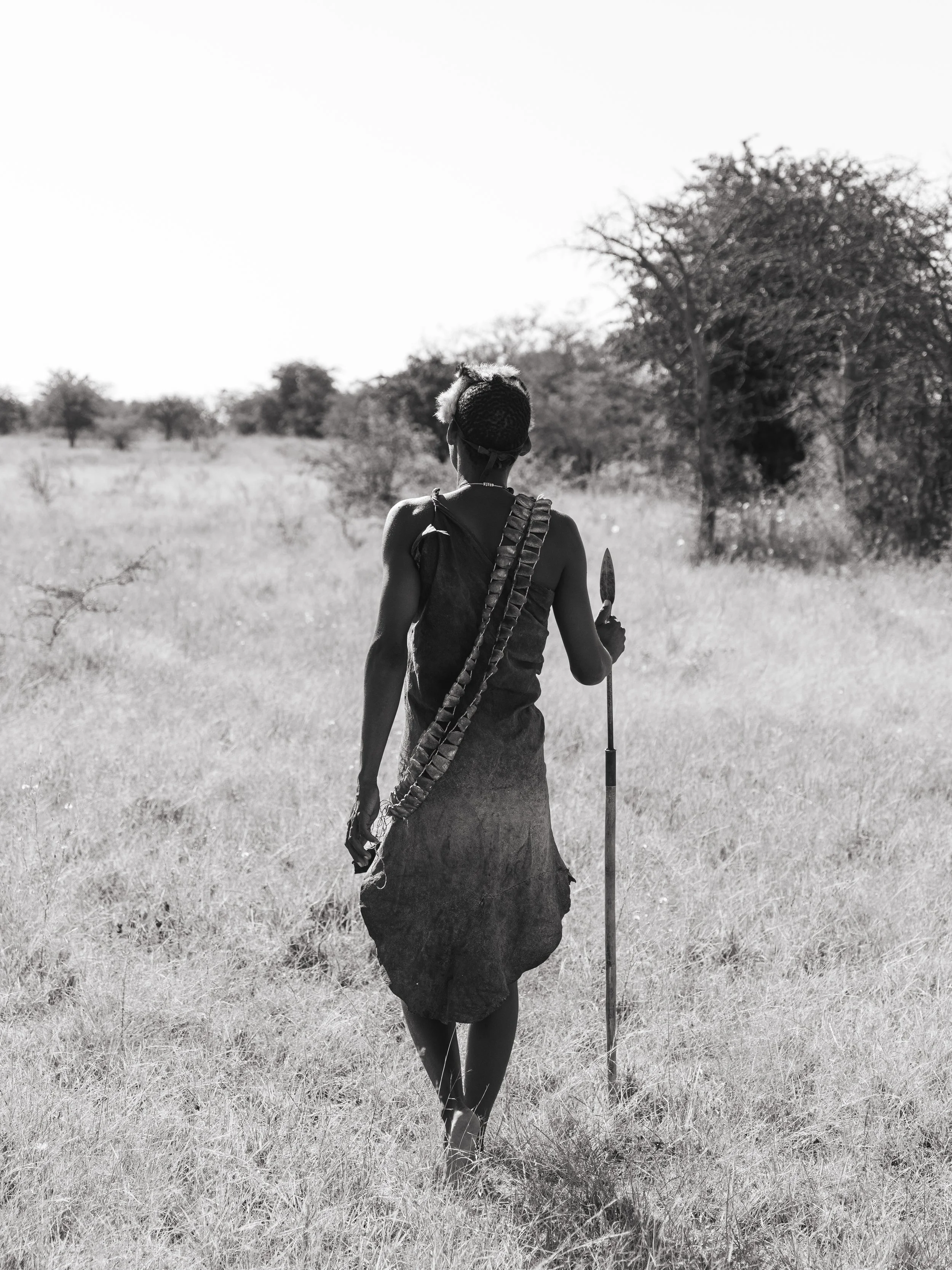 A woman with decorated hair, wearing a dress, standing in a field holding a spear, facing away from the camera, with trees in the background.