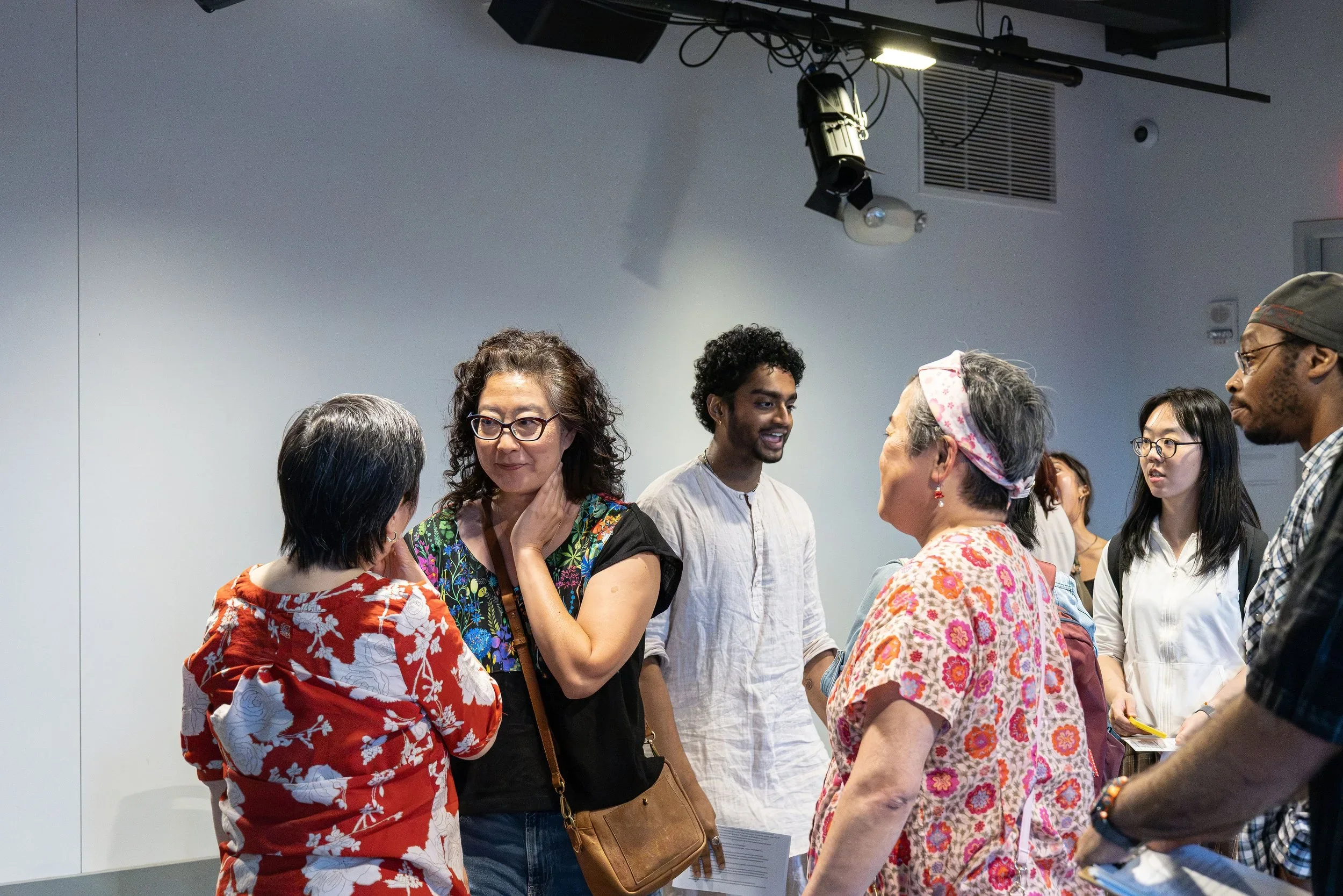 Group of diverse people engaged in conversation at an indoor event, with a plain light gray wall in the background.
