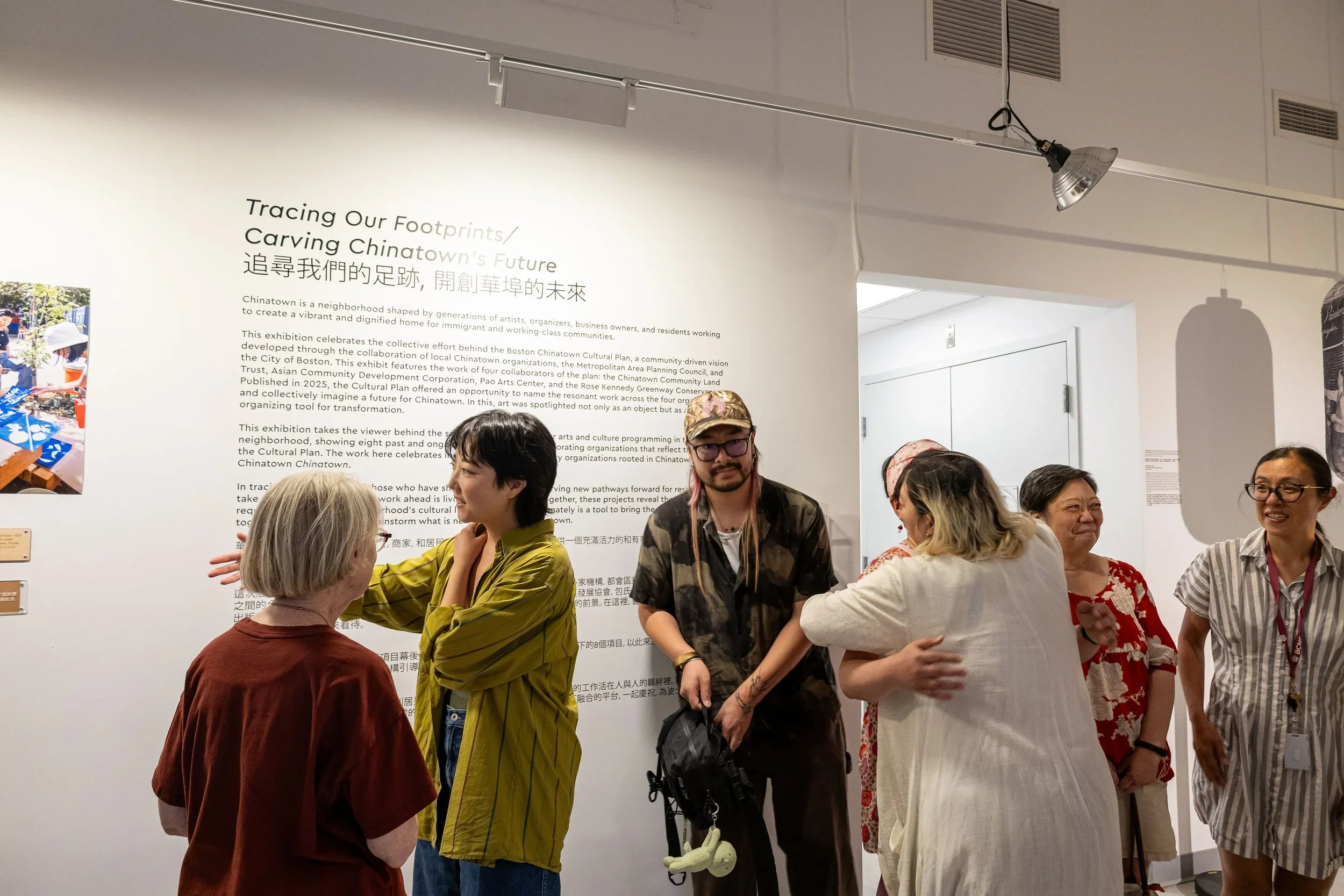A group of diverse people interacting and talking at an indoor exhibition or event, with a large display about Chinatown and its future on the wall behind them.