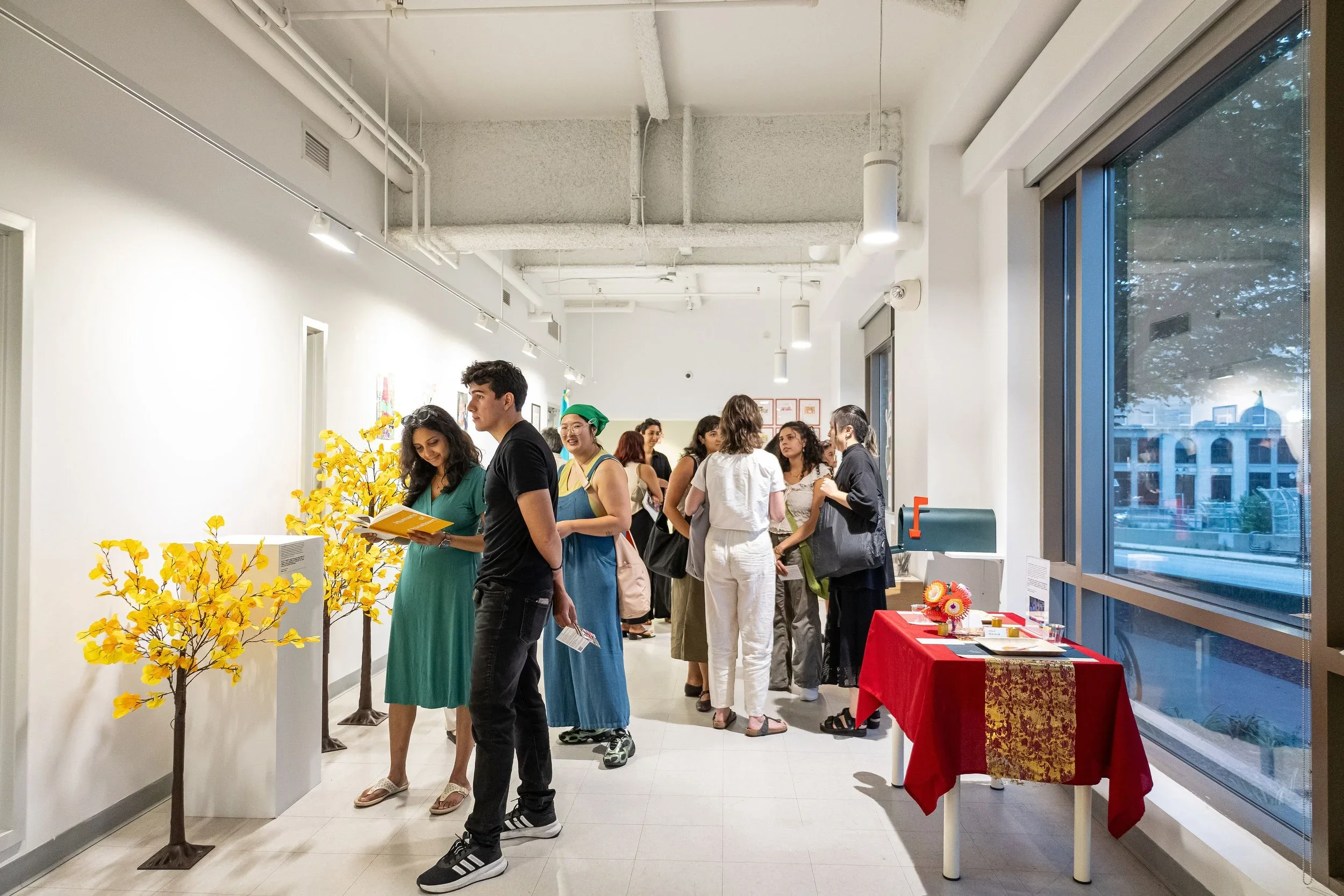 People attending an art or cultural exhibition in a gallery with artwork, flowers, and display tables, with a large window showing city buildings outside.