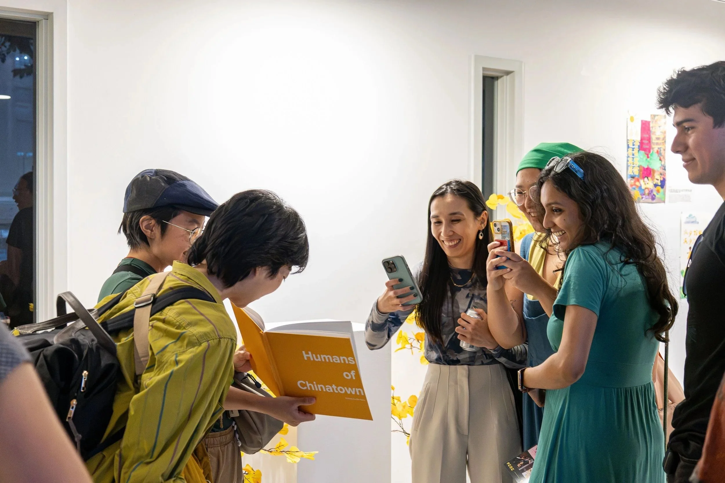 A group of diverse people gathered around a table looking at a book, smiling, in a well-lit room with white walls and some colorful decorations.